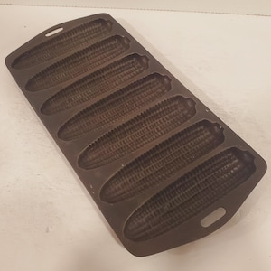 May include: A cast iron corn on the cob baking pan with six individual moulds. The pan is dark grey and has a handle on the side.