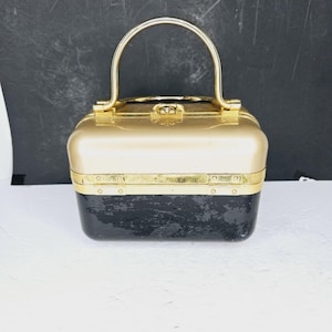 Può includere: Una borsa vintage oro e nera con manico dorato. La borsa rettangolare ha la parte superiore dorata e la parte inferiore nera. Il manico è in metallo dorato curvo. La borsa ha una chiusura a scatto.