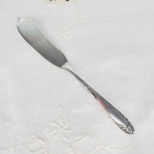 Può includere: Un coltello da burro d'argento con un designo ornamentale sul manico. Il coltello è su una tovaglia bianca con un disegno ricamato.