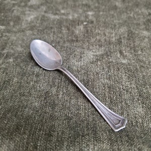 Puede incluir: Una cucharita de plata con un cuenco redondeado y un diseño simple y elegante. La cuchara tiene un mango ligeramente adornado con un patrón sutil.