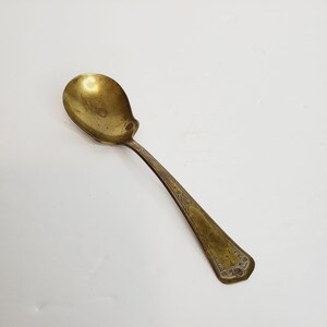 May include: A gold-toned spoon with an ornate design on the handle. The spoon has a rounded bowl and a long, slender handle.