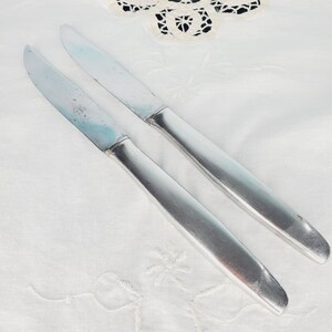 May include: Two silver butter knives with slightly rounded tips. The knives are lying on a white tablecloth with a lace pattern.