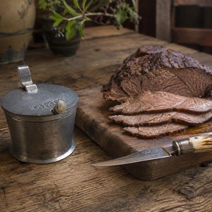 May include: A metallic lidded mustard pot with the letters JS and a symbol engraved on the lid, positioned next to a wooden cutting board holding a sliced cooked roast beef and a knife with a textured handle featuring a brown and white pattern.