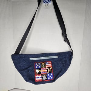 May include: A denim fanny pack with a black adjustable strap and a zipper closure. The pack features a patch with American flag imagery, an eagle, and the words "Proud American."