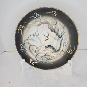 May include: A black and white ceramic plate with a gold rim features a detailed illustration of a dragon in flight. The dragon is painted in shades of gray and white, with accents of blue and gold. The plate has a black background with a white center.