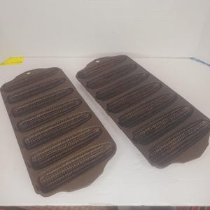May include: Two cast iron corn-shaped baking pans. Each pan has six corn cob moulds. The pans are dark brown and have a textured surface. The pans have a small hole at the top for hanging.