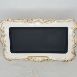 May include: White ornate frame with a black chalkboard insert and a gold metal stand. The frame has a distressed look with gold accents.