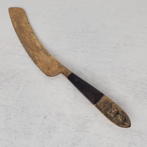 May include: A brass letter opener with a wooden handle. The handle is decorated with a carved design. The blade is curved and has a dull finish.