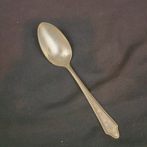 May include: A vintage silver spoon with a tarnished finish. The spoon has an oval bowl and a long handle with an ornate, engraved design. The handle's end features a decorative, geometric pattern.
