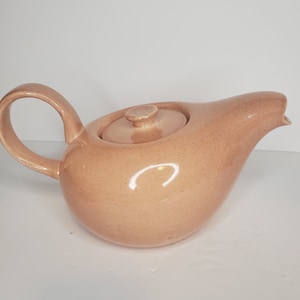 Puede incluir: Una tetera de cerámica rosa claro con cuerpo redondeado y un pico. La tetera tiene una tapa con un pomo en la parte superior.