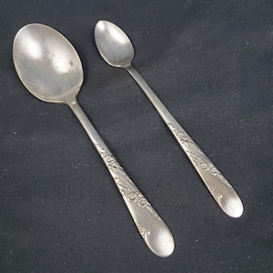 May include: Two silver spoons with ornate floral designs. The larger spoon is slightly curved and the smaller spoon is straight.