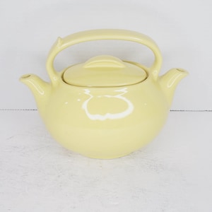 May include: A pale yellow ceramic teapot with a rounded body and a curved handle. The teapot has a lid with a small knob on top.
