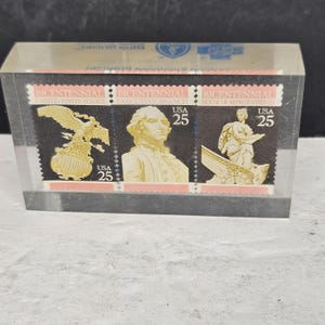 Puede incluir: Un juego de tres sellos conmemorativos del Bicentenario, cada uno con un valor de 25 centavos, exhibidos en una caja de plástico transparente. Los sellos presentan imágenes de un águila, George Washington y una figura clásica. Los sellos son negros, dorados y rosas.