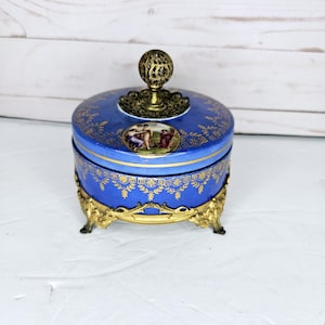 Puede incluir: Un joyero redondo de porcelana azul con patrones de filigrana dorada y un medallón central que representa figuras. La tapa está rematada con un asa esférica de latón y la base se apoya sobre cuatro patas decorativas de latón.
