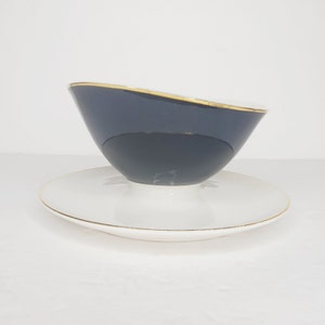 May include: A black and white ceramic teacup and saucer set with a gold trim. The teacup is tilted and the saucer is underneath.