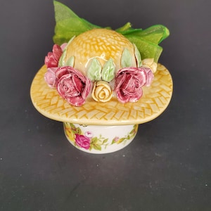 May include: A ceramic trinket box shaped like a straw hat with a floral design. The hat is decorated with pink, yellow, and green flowers. The box is white with a floral pattern.