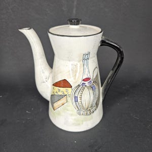 Puede incluir: Una tetera de cerámica blanca con asa y tapa negras. La tetera está decorada con ilustraciones de queso y una botella de vino. El pico es largo y curvado. La tetera probablemente se utiliza para servir bebidas.