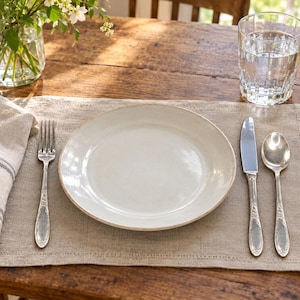 May include: A beige linen placemat featuring a round off-white ceramic plate centered on it. To the left is a silver fork, and to the right is a silver knife and spoon. A folded linen napkin with grey stripes sits beside the fork.