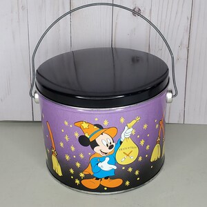 Puede incluir: Lata de metal morada para golosinas de Halloween con tapa negra y asa. La lata presenta una imagen de dibujos animados de Mickey Mouse vestido de mago sosteniendo una bolsa que dice "Trick or Treat".