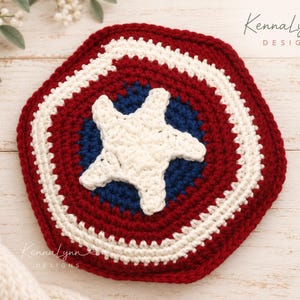 May include: A round, crocheted textile piece featuring a red, white, and blue circular pattern with a large white star in the centre. The design resembles a shield. The text KennaLynn DESIGNS is visible in the top right and bottom left corners.