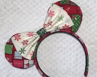 May include: A headband featuring two circular ears and a central bow. The ears are covered in a red fabric with a pattern of colorful gift boxes. The bow is made of white fabric with red and green snowflake prints and a green center knot with the word Joy.