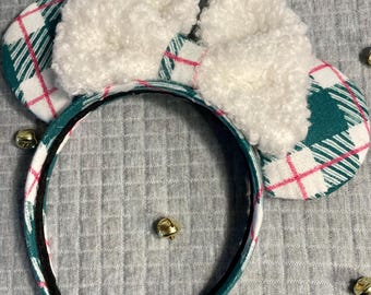 May include: A festive headband with plaid ears in green and white, accented with pink lines. The ears are topped with fluffy white bows. Gold bells are scattered around the headband, suggesting a holiday theme.