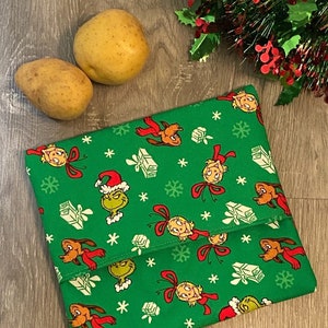 May include: Green fabric lunch bag with a repeating pattern of the Grinch, Cindy Lou Who, and Max the dog. The bag is folded in half and has a green background with white snowflakes and presents. The text "Green Grinch" is printed on the bottom of the image.