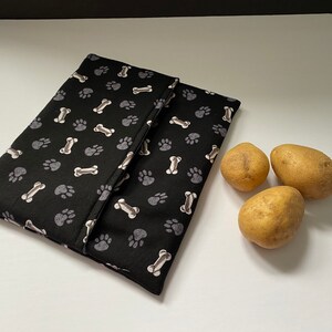 Puede incluir: Una bolsa de tela negra con un patrón de huellas de patas y huesos. La bolsa está doblada por la mitad y está sobre una superficie blanca. Hay tres patatas al lado de la bolsa.