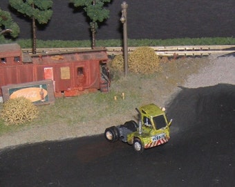 May include: A miniature model of a green and yellow semi-truck with a trailer. The truck is parked in front of a red and brown boxcar with a Coca-Cola advertisement on the side.