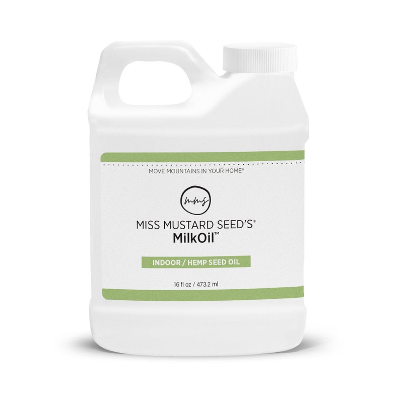 May include: A white plastic container of Miss Mustard Seed's MilkOil. The container has a handle and a white screw-on cap. The label includes the text "MOVE MOUNTAINS IN YOUR HOME", "INDOOR / HEMP SEED OIL", and "16 fl oz / 473.2 ml".