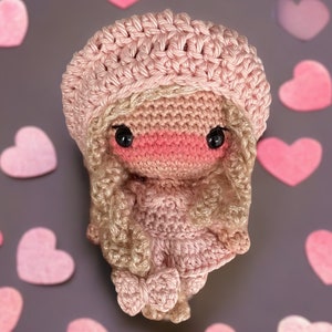Pu&ograve; includere: Una bambola all'uncinetto realizzata con filato rosa e marrone, che indossa un cappello rosa e un vestito rosa. La bambola ha occhi marroni e un naso rosa.