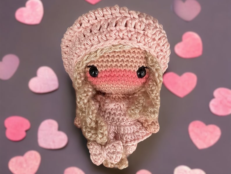 Pu&ograve; includere: Una bambola all'uncinetto realizzata con filato rosa e marrone, che indossa un cappello rosa e un vestito rosa. La bambola ha occhi marroni e un naso rosa.