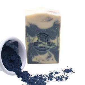 May include: A bar of soap with a marbled design in shades of cream and dark blue, with the text "Wash Up Balancer Soap" imprinted on the front. A small white bowl spills dark blue powder onto the white surface.