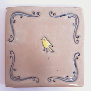 Puede incluir: Un azulejo de cerámica cuadrado con fondo marrón y un borde decorativo negro y azul. Un pájaro amarillo está pintado en el centro del azulejo.