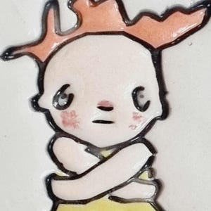 Puede incluir: Una figurita de cerámica pintada a mano de una niña con el pelo naranja, que lleva un vestido amarillo y tiene los brazos cruzados.