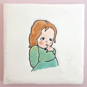 May include: A square ceramic tile featuring a hand-drawn illustration of a young child. The child has red hair, rosy cheeks, and is wearing a teal dress with yellow polka dots. The child is touching their finger to their mouth.