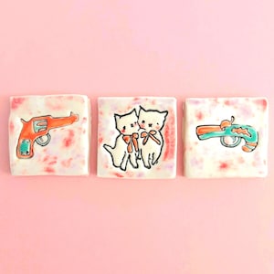 May include: Three square ceramic tiles with a mottled pink and white background. The left tile features an orange revolver, the centre tile shows two white cats with orange bows, and the right tile displays a teal and orange toy water pistol.