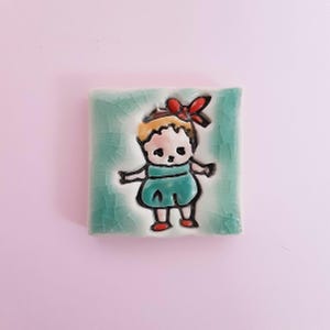 Puede incluir: Un azulejo de cerámica con una ilustración pintada a mano de una niña con cabello rubio, que lleva un vestido azul y un lazo rojo. El azulejo tiene un fondo verde.