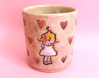 May include: A cylindrical ceramic mug with a glossy, mottled pink and beige glaze. The exterior features a hand-painted illustration of a small figure with yellow hair and a white dress, surrounded by multiple small, red heart-shaped motifs.
