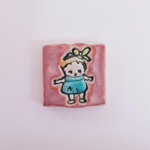 Può includere: Una piastrella di ceramica rosa con un'illustrazione dipinta a mano di una ragazza con un fiocco giallo e un vestito blu. La ragazza sorride e ha le braccia distese.