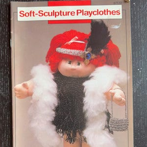 May include: A vintage doll in a flapper outfit, featuring a black dress, white boa, and a beaded purse. The doll has red hair and a jeweled headband. The text "Soft-Sculpture Playclothes" is at the top of the image.