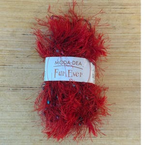 Pode incluir: Uma bola de lã vermelha Moda Dea Fur Ever. O fio tem uma textura difusa com fios vermelhos e pretos. A etiqueta no fio é branca com as palavras "Moda Dea" e "Fur Ever" em fonte preta.
