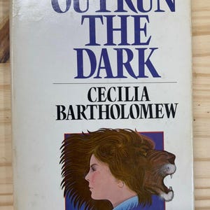 May include: A paperback book titled "Outrun the Dark" by Cecilia Bartholomew. The cover features a woman's profile with a lion's head, set against a blue and white background. The book's title is in large, dark blue letters.
