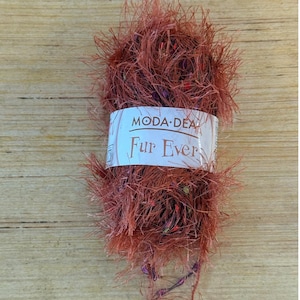Pode incluir: Uma bola de fio castanho-avermelhado, semelhante a pele, com o texto "Fur Ever" numa etiqueta branca. O fio tem um aspeto texturizado e fofo, com fios de comprimentos variados. A etiqueta também inclui o texto "MODA-DEA".