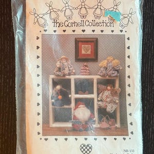 May include: A package featuring a display of handmade clothespin dolls. The Cornell Collection logo is at the top. The dolls are arranged on a shelf, with a Santa doll at the bottom. The package is sealed in a clear plastic bag.