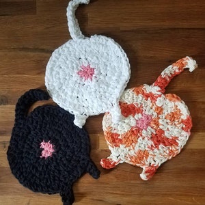 May include: Three crocheted cat-shaped dishcloths. One is white, one is black, and one is orange and white.