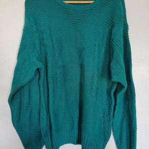 Puede incluir: Suéter de color verde azulado con cuello redondo y mangas largas. El suéter presenta un diseño de punto de cable texturizado y puños y dobladillo acanalados. La etiqueta dice "Jantzen". El suéter está colgado en una percha de madera.