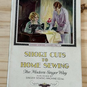 May include: A vintage booklet titled "Short Cuts to Home Sewing The Modern Singer Way" published by Singer Sewing Machine Co. Inc. The cover features an illustration of two women sewing on a Singer sewing machine.