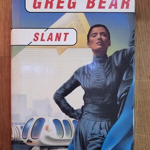 May include: A book cover with a blue and white futuristic cityscape background. The title "SLANT" is in white letters on a red rectangle. The author's name "GREG BEAR" is in red letters on a white rectangle. A woman in a blue jumpsuit and a blue cape is standing in front of a white futuristic vehicle.