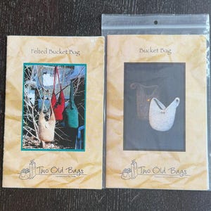 May include: Two patterns for felted bucket bags. One pattern shows a beige, red, and green felted bucket bag hanging from a tree branch. The other pattern shows two felted bucket bags in gray and white. The text on the patterns reads "Felted Bucket Bag" and "Bucket Bag".